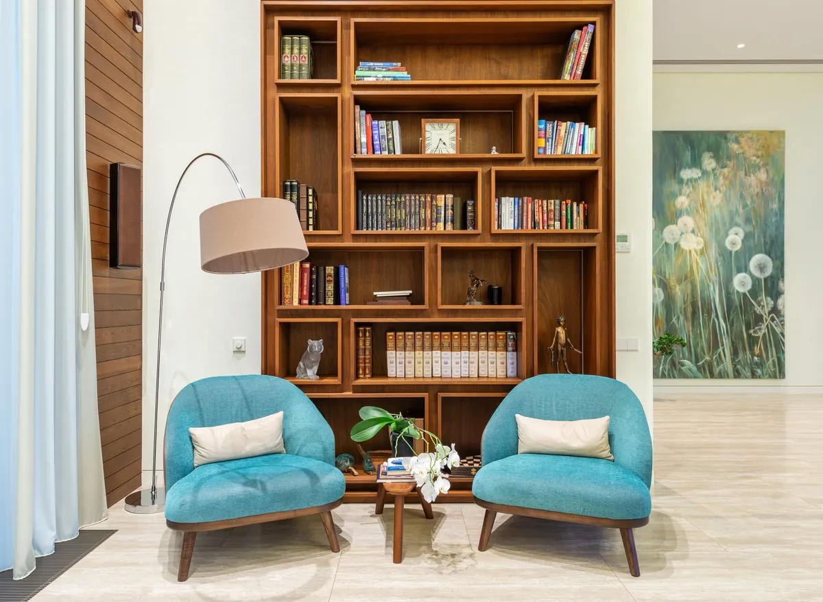Une bibliothèque en bois remplie de livres, devant deux fauteuils bleus et un lampadaire